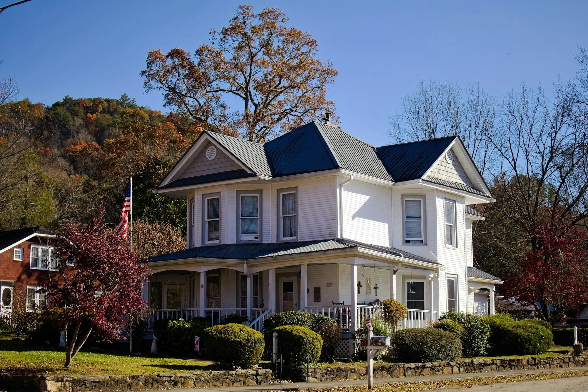 White Victorian home with a dark shingle roof and wraparound porch in fall